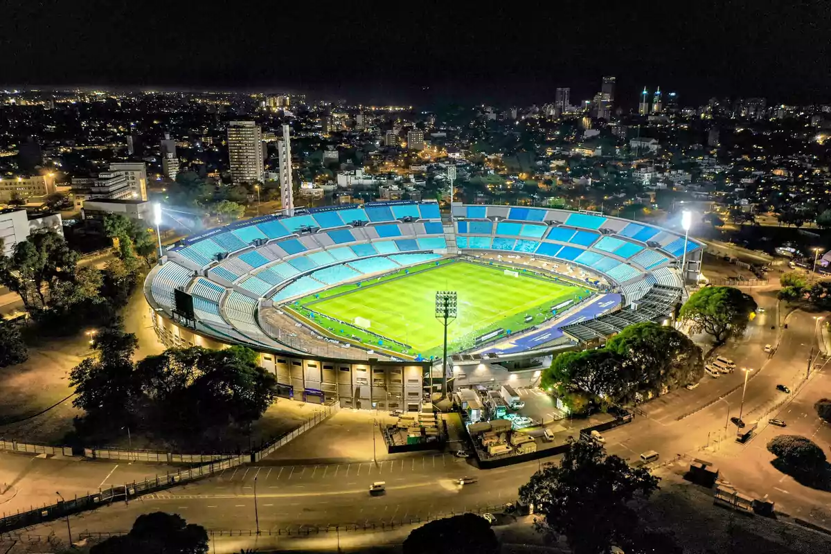 Vista aérea nocturna de un estadio de fútbol iluminado con gradas celestes y la ciudad al fondo Vista aérea nocturna de un estadio de fútbol iluminado con gradas celestes y la ciudad al fondo