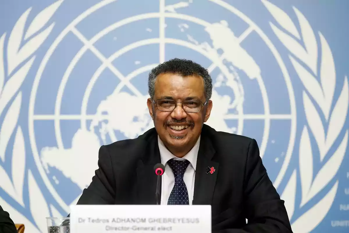 Hombre con gafas y traje oscuro sonríe mientras está sentado frente a un micrófono, con un fondo azul que muestra el logotipo de las Naciones Unidas.