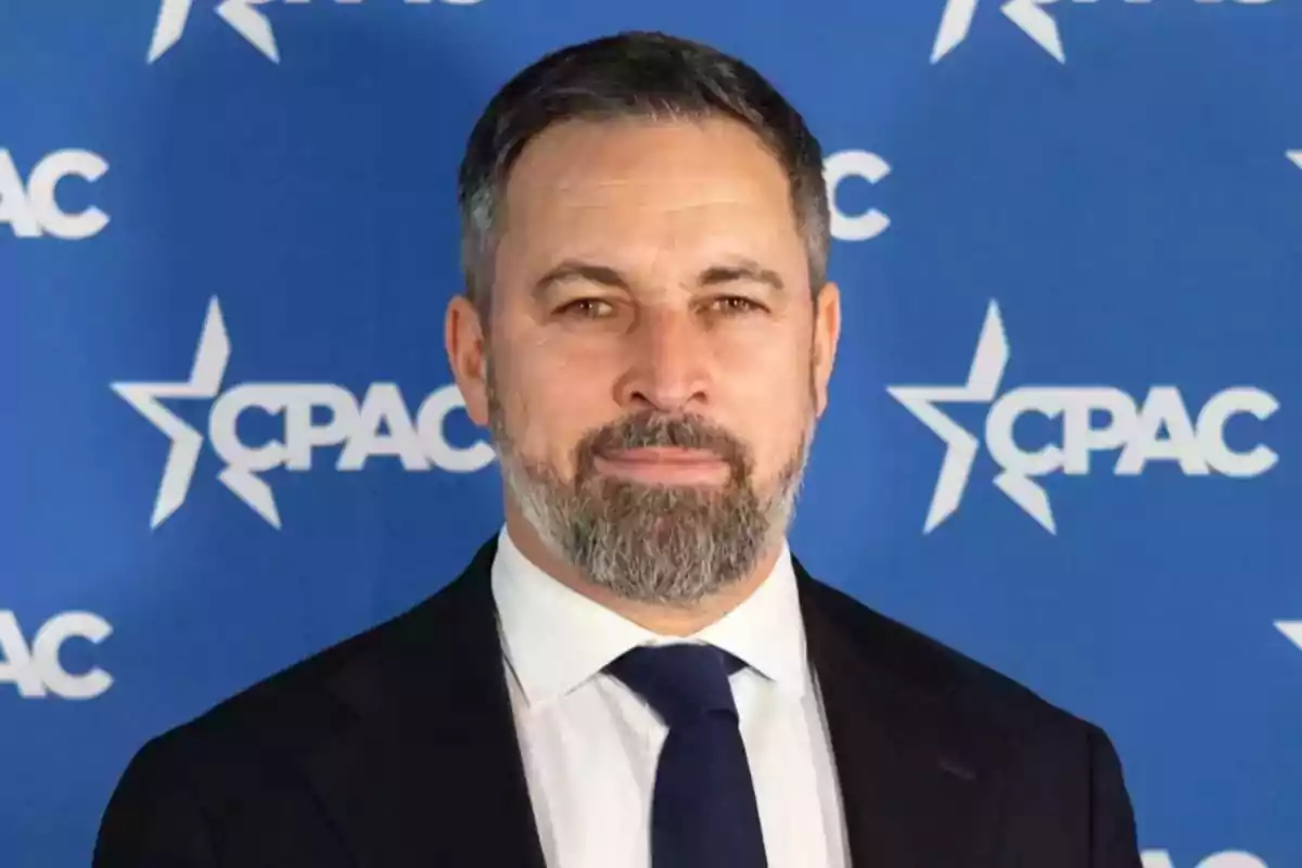 Short-haired, bearded man in a suit and tie posing in front of a blue background with the CPAC logo repeated.