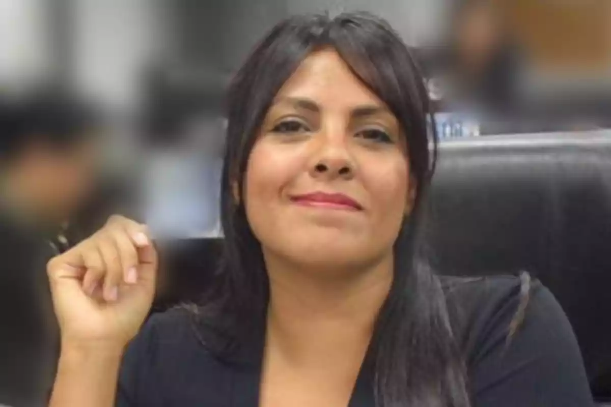 Mujer de cabello oscuro y lacio sentada en una silla negra, con expresión sonriente y fondo desenfocado. Mujer de cabello oscuro y lacio sentada en una silla negra, con expresión sonriente y fondo desenfocado.