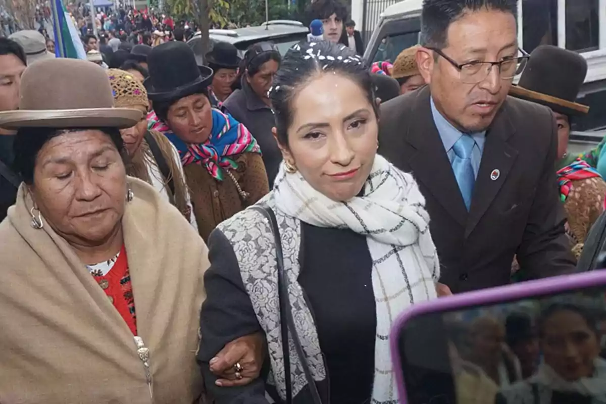 A woman wearing a white scarf and a dark jacket walks accompanied by other people in a crowd, some of them dressed in traditional Andean clothing and hats, while in the background a street full of people can be seen. A woman wearing a white scarf and a dark jacket walks accompanied by other people in a crowd, some of them dressed in traditional Andean clothing and hats, while in the background a street full of people can be seen.