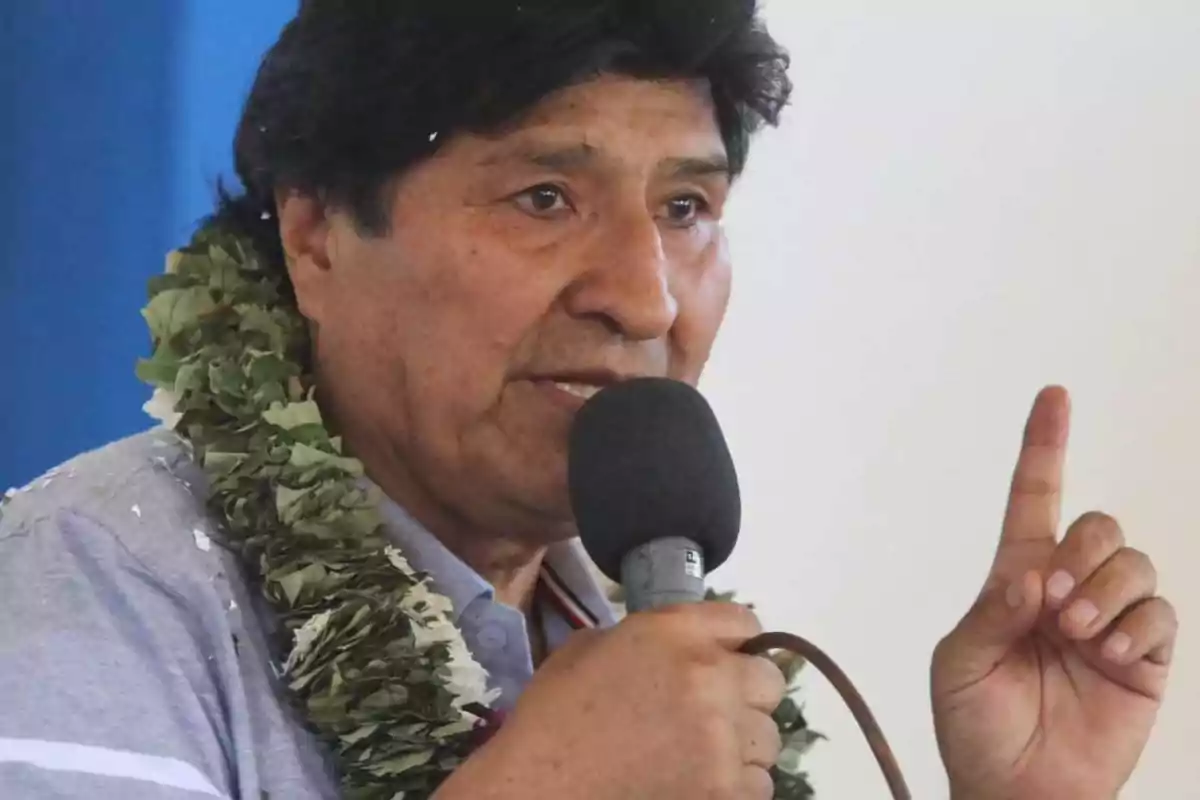 Hombre de cabello oscuro con guirnalda de hojas hablando por micrófono y levantando un dedo Hombre de cabello oscuro con guirnalda de hojas hablando por micrófono y levantando un dedo