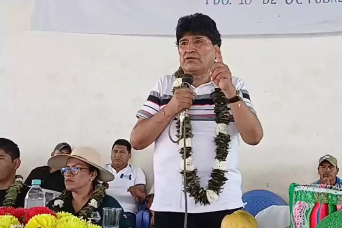 Un hombre de pie con micrófono y guirnalda de flores habla en un evento mientras varias personas lo observan sentadas a su alrededor