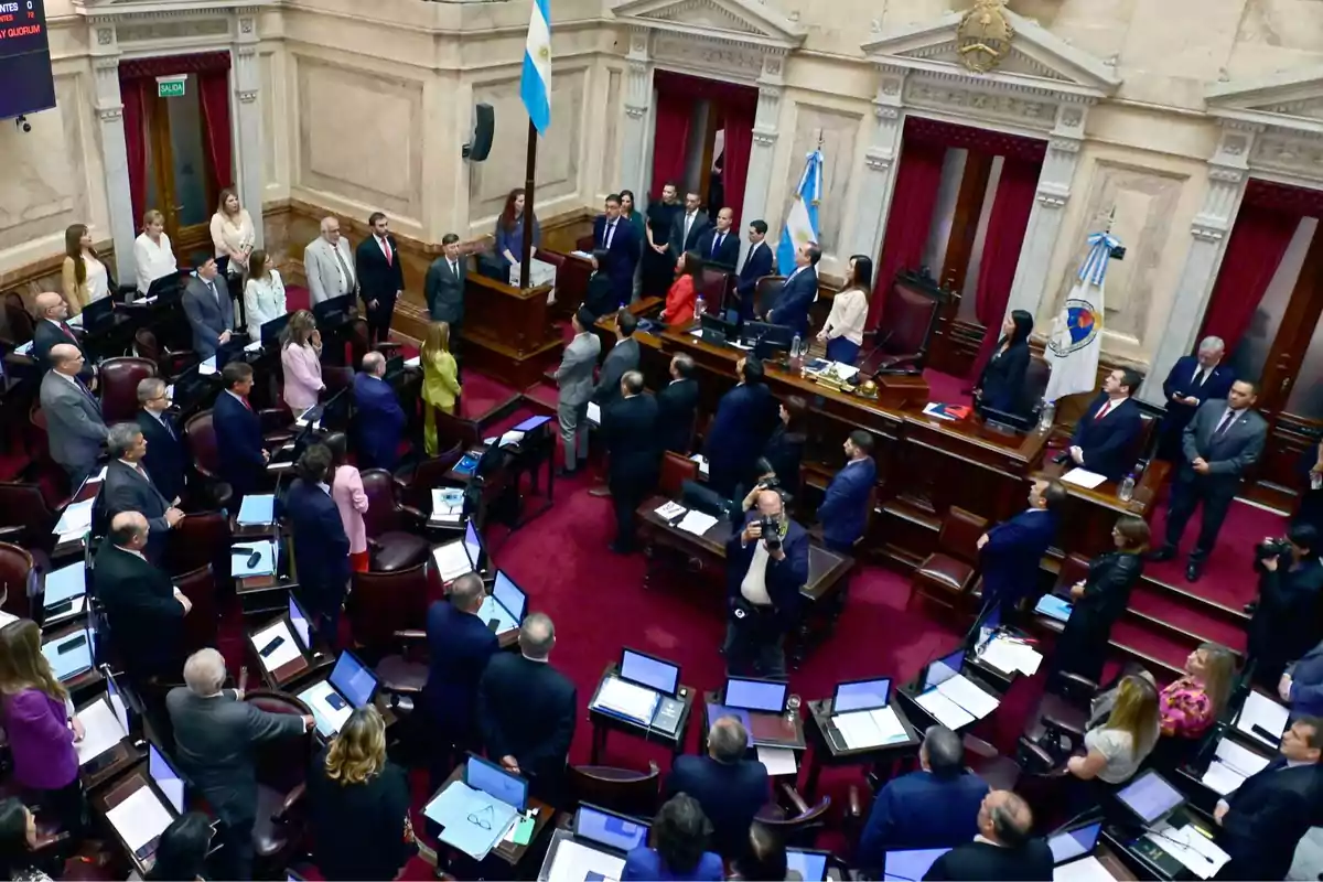 Overview of the Argentine Senate chamber with legislators and officials standing during a session, surrounded by desks with computers and national flags in a solemn atmosphere