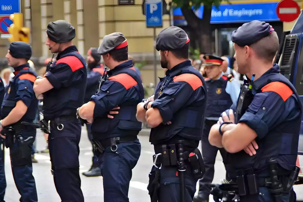 Varios policías uniformados con boinas grises y detalles rojos están de pie en fila con los brazos cruzados en una calle urbana.