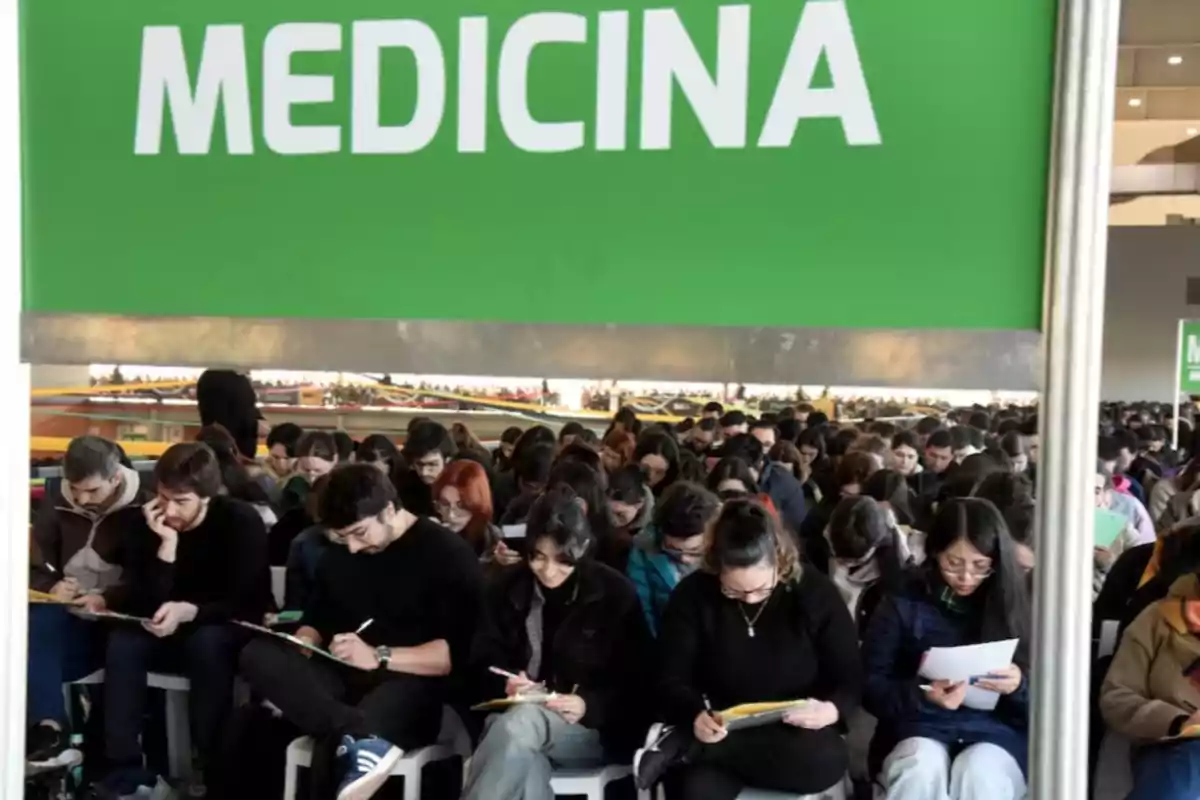 Un gran grupo de estudiantes sentados tomando apuntes o exámenes bajo un cartel verde que dice MEDICINA Un gran grupo de estudiantes sentados tomando apuntes o exámenes bajo un cartel verde que dice MEDICINA