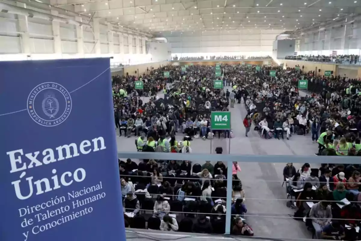 Miles de personas sentadas en un gran salón cerrado rindiendo un examen, con carteles verdes que indican diferentes áreas y un cartel azul del Ministerio de Salud de Argentina en primer plano.