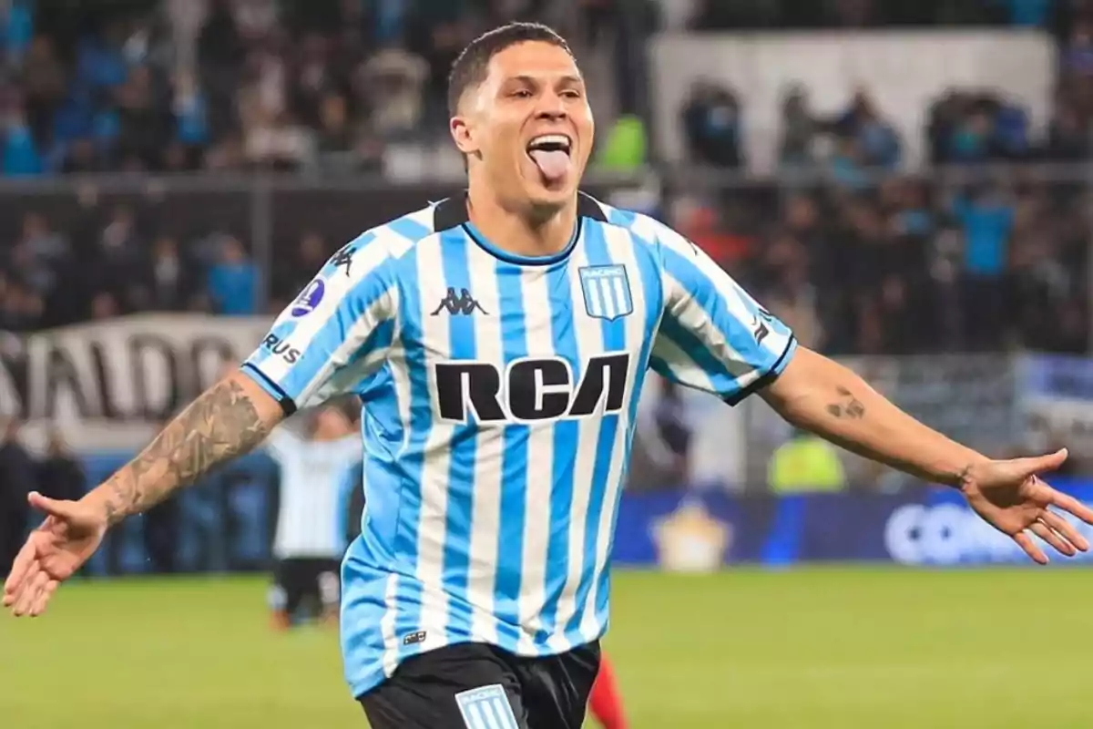 Soccer player celebrating a goal with arms outstretched and tongue out, wearing the Racing Club jersey in a stadium full of fans Soccer player celebrating a goal with arms outstretched and tongue out, wearing the Racing Club jersey in a stadium full of fans