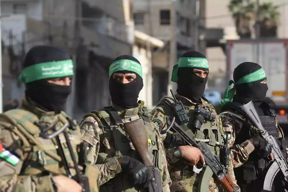 Cuatro hombres armados con uniformes de camuflaje y pasamontañas negros, portando armas y bandas verdes en la cabeza, están de pie en una calle urbana