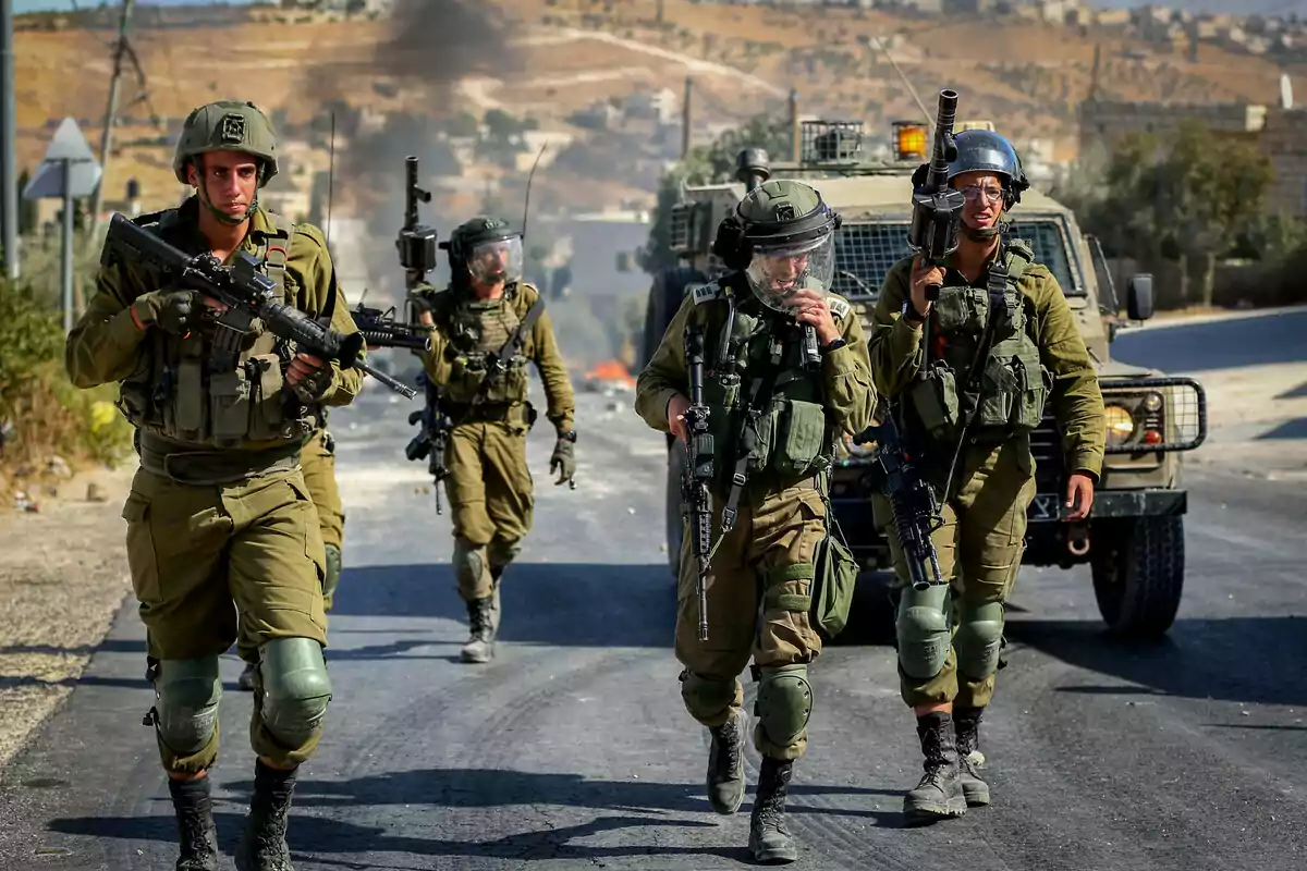 Soldados armados caminan por una carretera junto a un vehículo militar en una zona árida con humo al fondo Soldados armados caminan por una carretera junto a un vehículo militar en una zona árida con humo al fondo