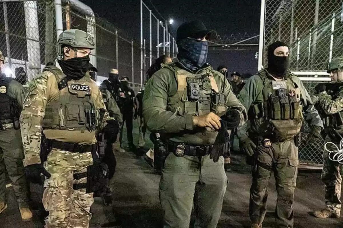 Group of heavily armed police officers with tactical gear and balaclavas patrolling a fenced-off area at night