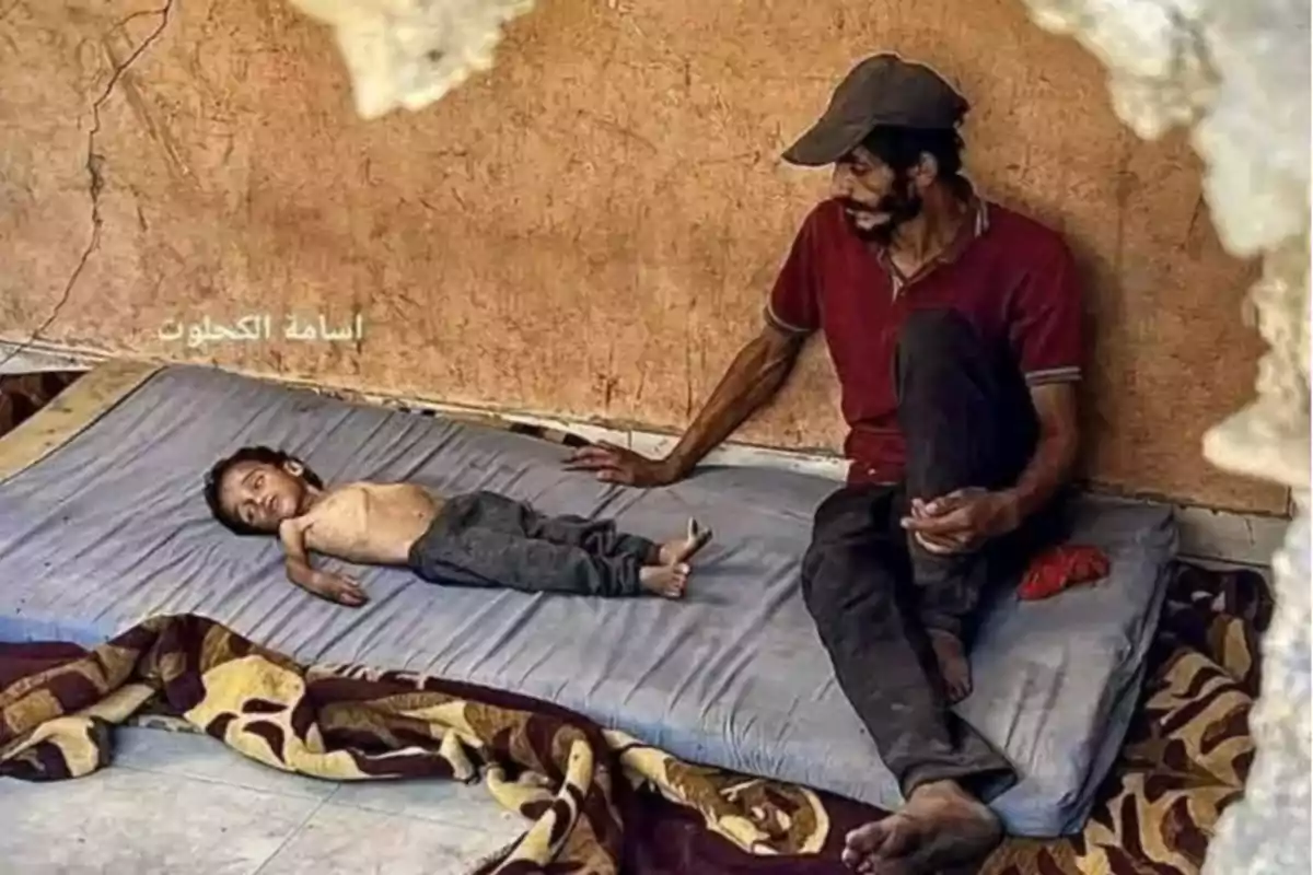 A man and a boy rest on a mattress on the floor inside a room with worn-out walls.