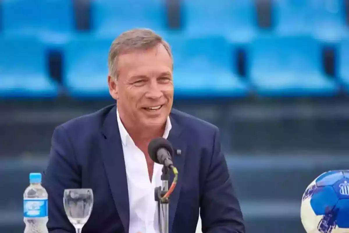 Hombre de traje azul sonriendo mientras habla frente a un micrófono en un estadio con asientos azules, con una botella de agua, una copa y un balón de fútbol sobre la mesa Hombre de traje azul sonriendo mientras habla frente a un micrófono en un estadio con asientos azules, con una botella de agua, una copa y un balón de fútbol sobre la mesa