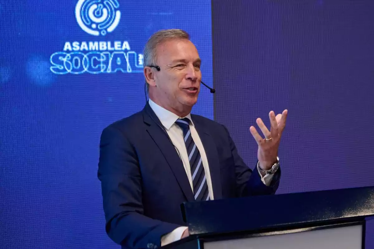 Hombre de traje hablando en un podio con micrófono de diadema frente a una pantalla azul que muestra el texto Asamblea Social Hombre de traje hablando en un podio con micrófono de diadema frente a una pantalla azul que muestra el texto Asamblea Social
