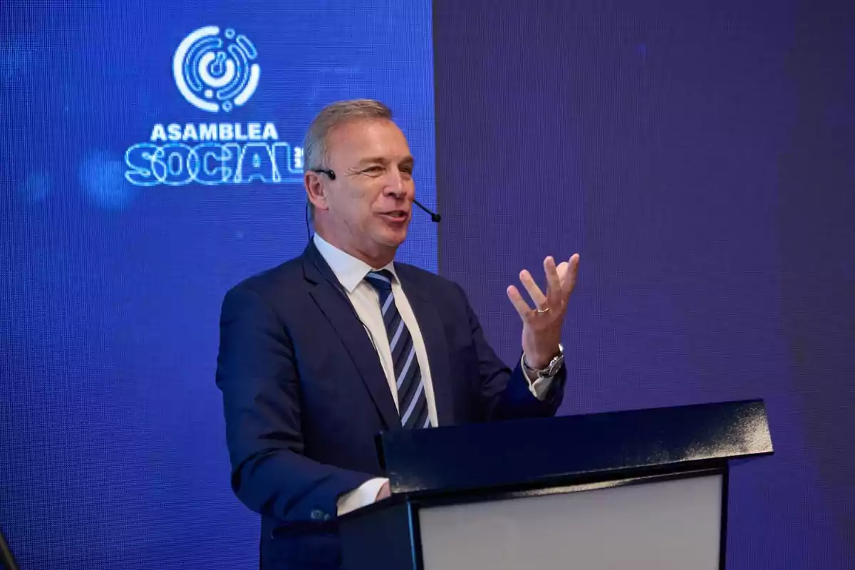 Hombre de traje hablando en un podio con micrófono de diadema y fondo azul que muestra el texto Asamblea Social