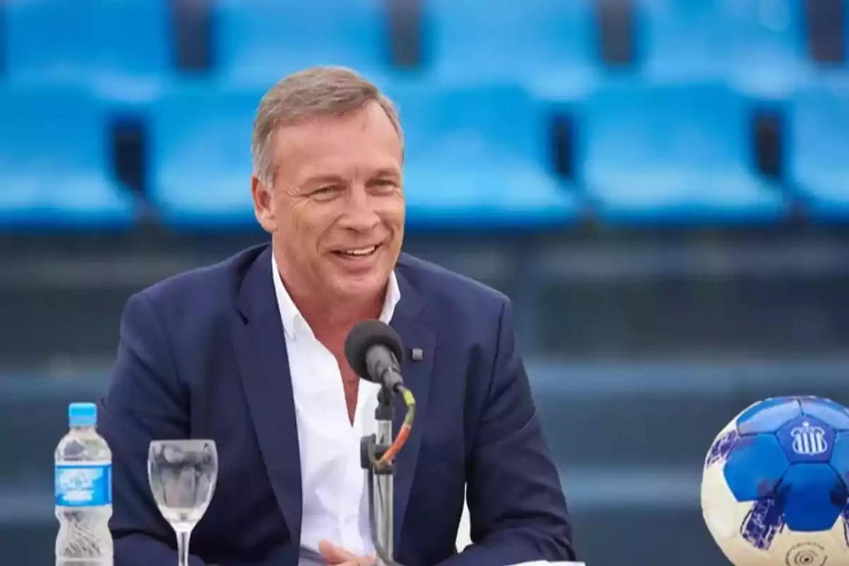 Hombre de traje azul sonriendo mientras habla frente a un micrófono en un estadio con asientos azules, con una botella de agua, una copa y un balón de fútbol sobre la mesa