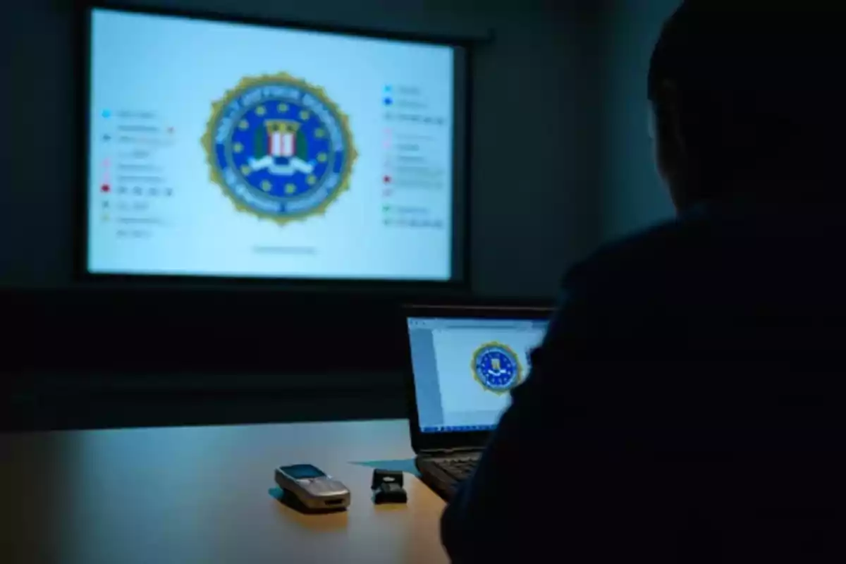 Person sitting in front of a laptop in a dark room with the FBI logo projected on a large screen in the background Person sitting in front of a laptop in a dark room with the FBI logo projected on a large screen in the background