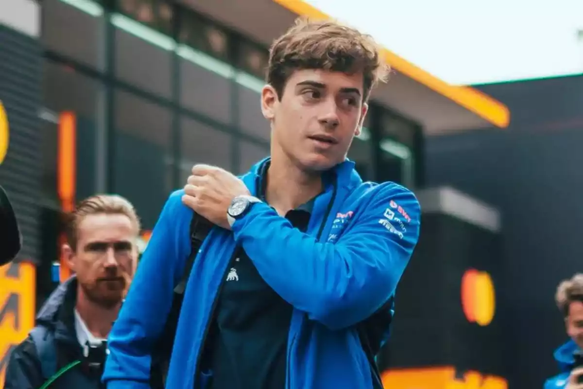 Joven con chaqueta azul y mochila caminando en un entorno urbano con personas al fondo