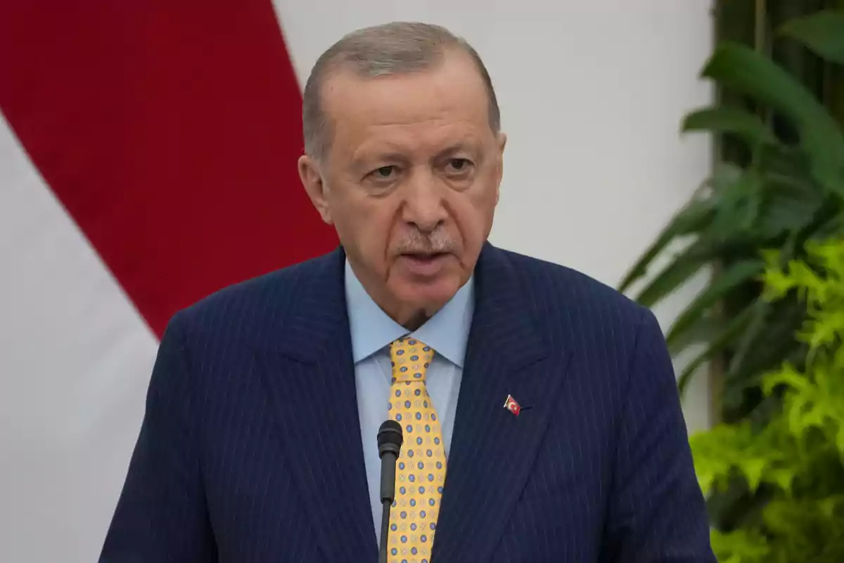 Man in a blue suit with a yellow tie speaking in front of a microphone with a background of plants and a red and white flag. Man in a blue suit with a yellow tie speaking in front of a microphone with a background of plants and a red and white flag.