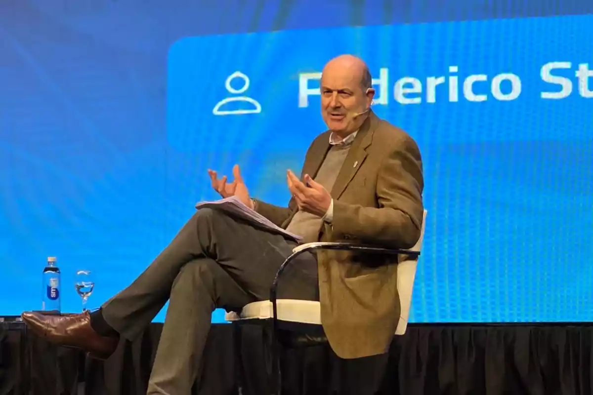 Man sitting on a chair on a stage with a blue background while speaking and holding some papers