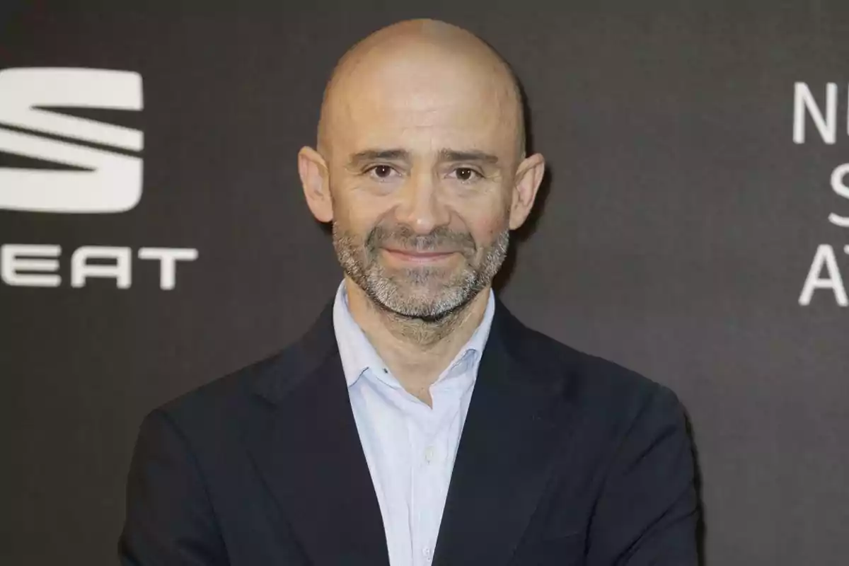 Bald man with a gray beard and a dark suit posing in front of a black background with white logos Bald man with a gray beard and a dark suit posing in front of a black background with white logos