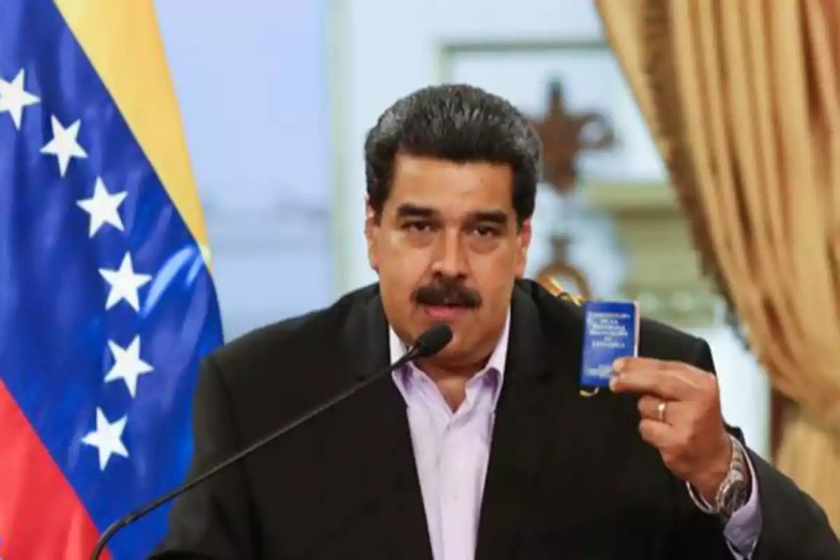 Hombre de cabello oscuro y bigote hablando frente a un micrófono mientras sostiene un pequeño libro azul con una bandera de Venezuela al fondo Hombre de cabello oscuro y bigote hablando frente a un micrófono mientras sostiene un pequeño libro azul con una bandera de Venezuela al fondo