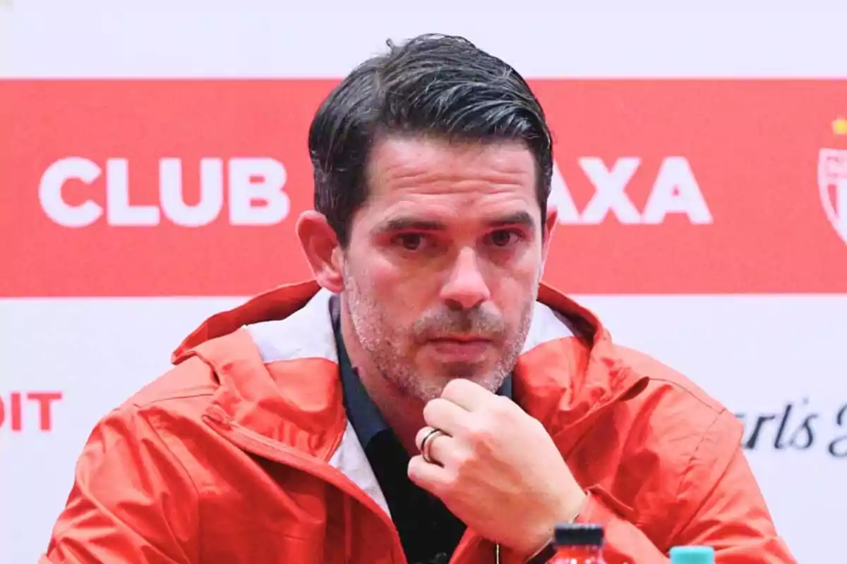 Man in a red jacket sitting in front of a background with white and red letters, resting his hand on his chin and looking straight ahead Man in a red jacket sitting in front of a background with white and red letters, resting his hand on his chin and looking straight ahead