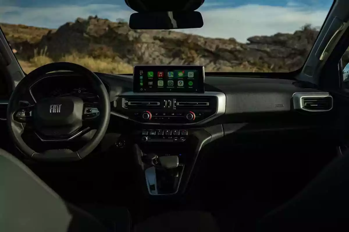 Interior de un automóvil Fiat moderno con volante, pantalla central encendida y controles de aire acondicionado, con paisaje rocoso visible a través del parabrisas