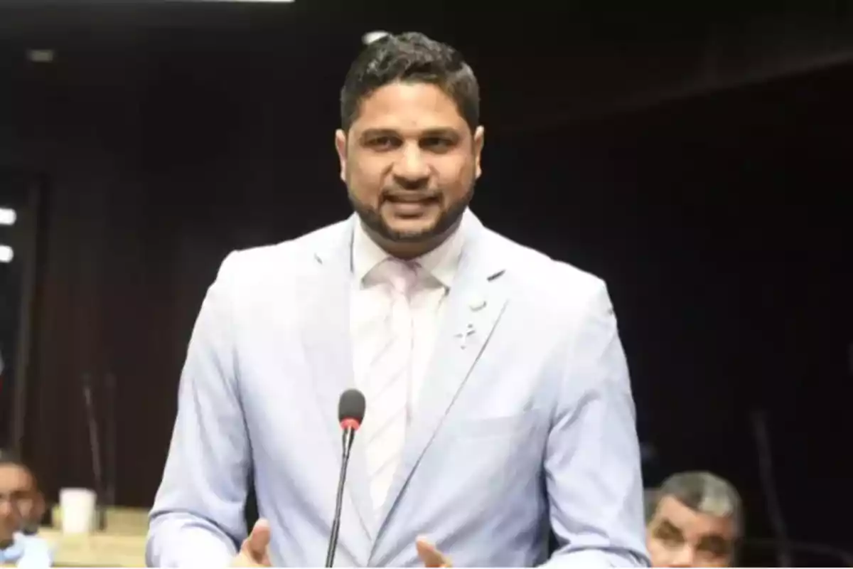 Hombre de pie hablando frente a un micrófono con saco claro y corbata en un entorno formal