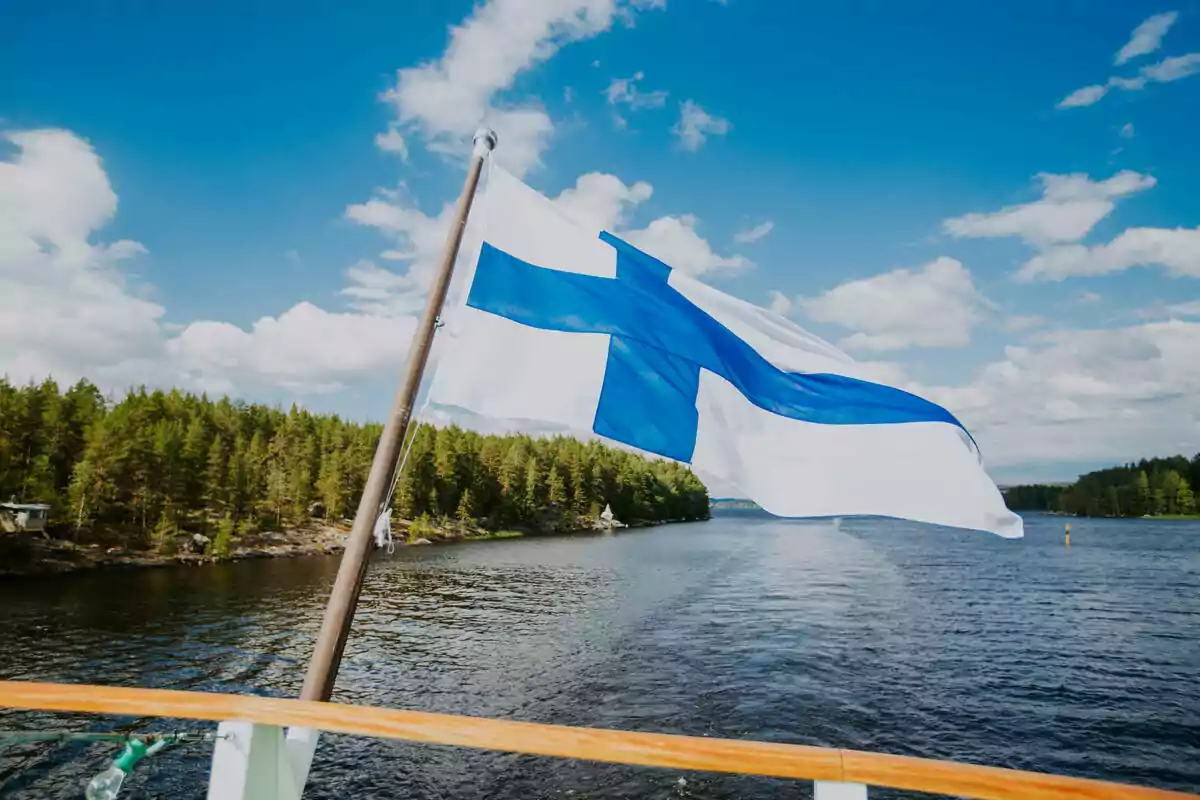 Bandera de Finlandia ondeando en un barco sobre un lago rodeado de árboles bajo un cielo azul con nubes