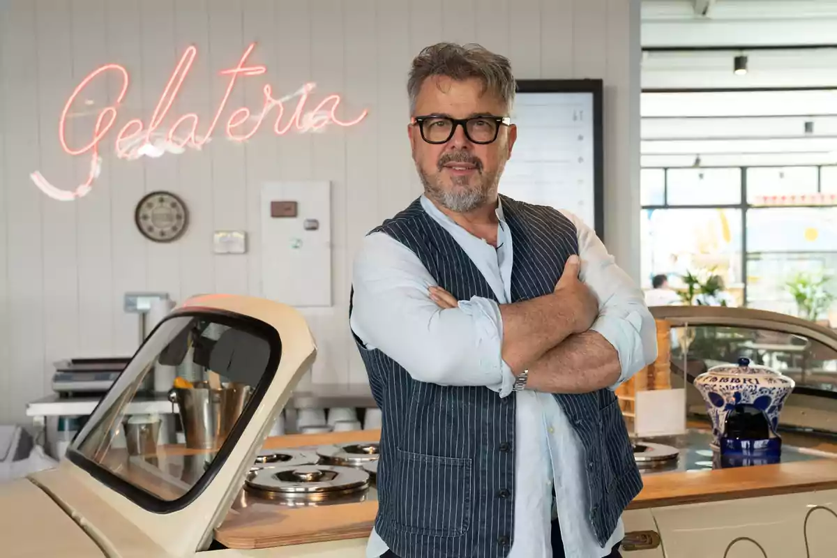 Gray-haired man with glasses standing with his arms crossed inside an ice cream shop next to an ice cream counter and a neon sign that says Gelateria