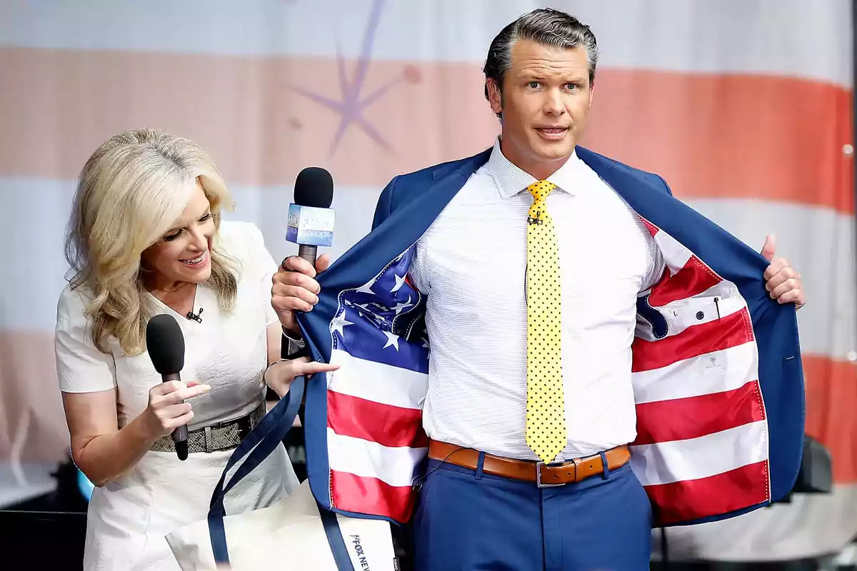 Un hombre con traje azul muestra el forro de su saco con diseño de la bandera de Estados Unidos mientras una mujer a su lado sonríe y sostiene un micrófono Un hombre con traje azul muestra el forro de su saco con diseño de la bandera de Estados Unidos mientras una mujer a su lado sonríe y sostiene un micrófono
