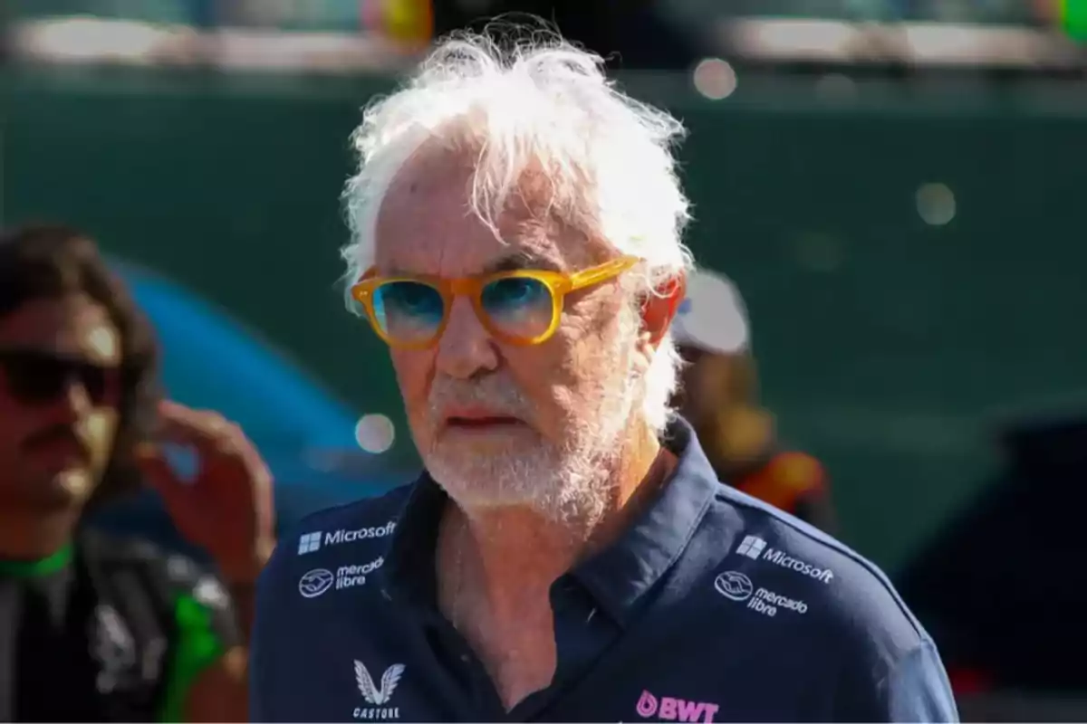 Gray-haired, gray-bearded man with yellow-framed glasses and blue lenses wearing a blue jersey with sponsor logos in an outdoor setting