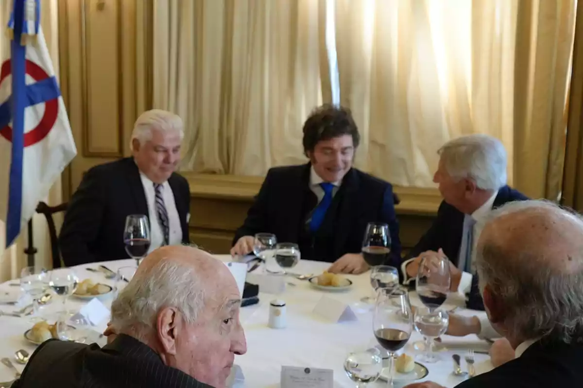 Five men in suits sit around an elegant table with wine glasses and bread, talking in a formal room. Five men in suits sit around an elegant table with wine glasses and bread, talking in a formal room.