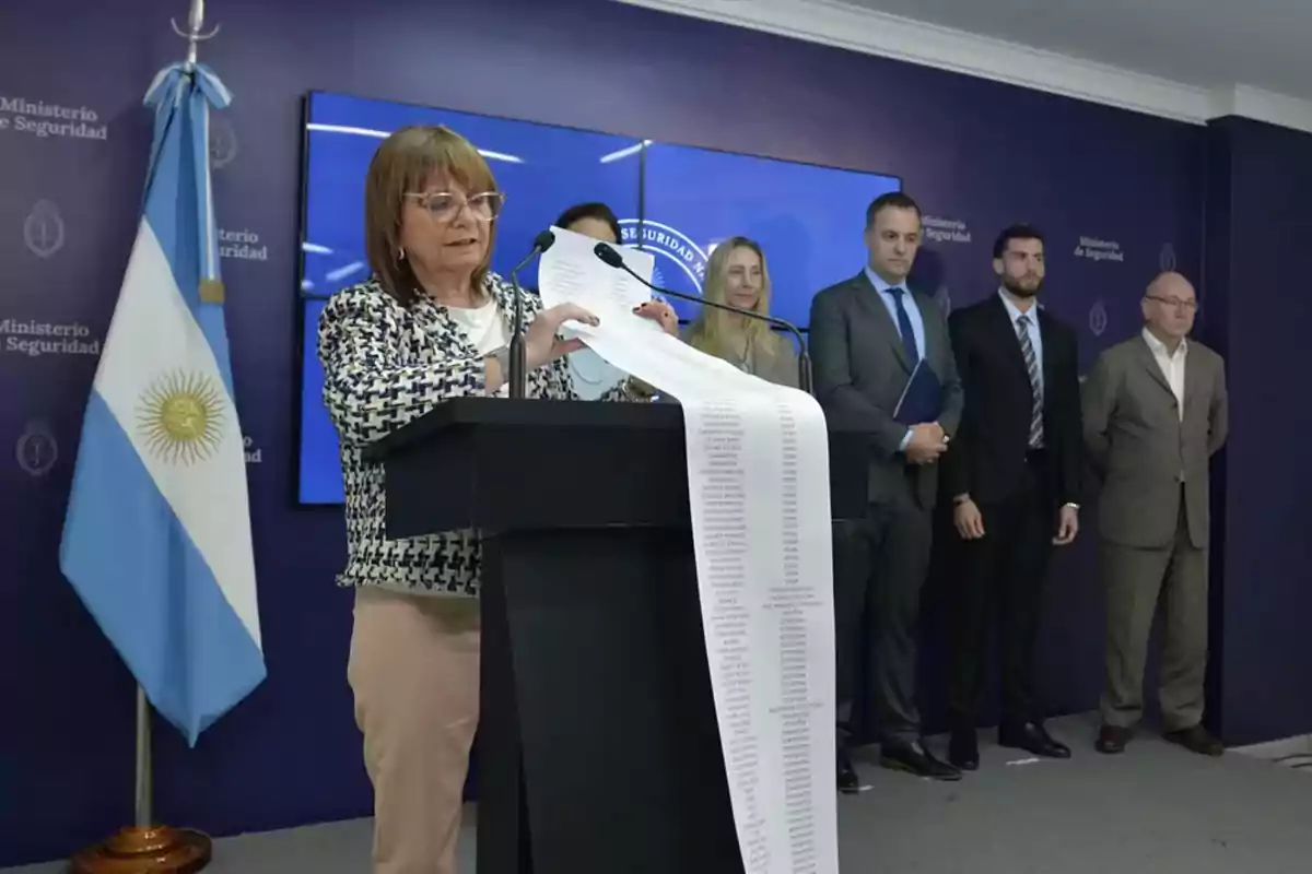 Una mujer de pie en un atril sostiene una larga lista de papel mientras varias personas la observan en una sala con fondo azul y una bandera de Argentina Una mujer de pie en un atril sostiene una larga lista de papel mientras varias personas la observan en una sala con fondo azul y una bandera de Argentina