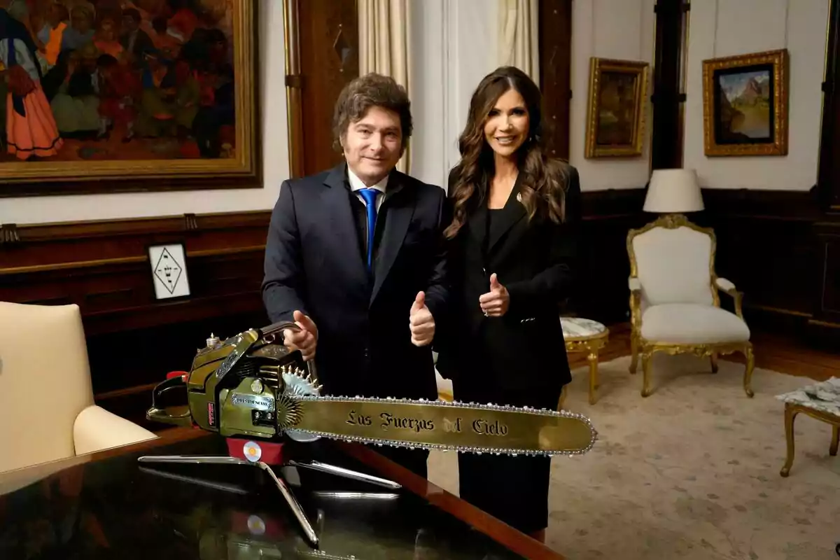 Dos personas de pie en una oficina elegante, ambas con traje oscuro y mostrando pulgares arriba, junto a una motosierra dorada sobre una mesa que tiene grabado el texto Los Fuerzas del Cielo Dos personas de pie en una oficina elegante, ambas con traje oscuro y mostrando pulgares arriba, junto a una motosierra dorada sobre una mesa que tiene grabado el texto Los Fuerzas del Cielo