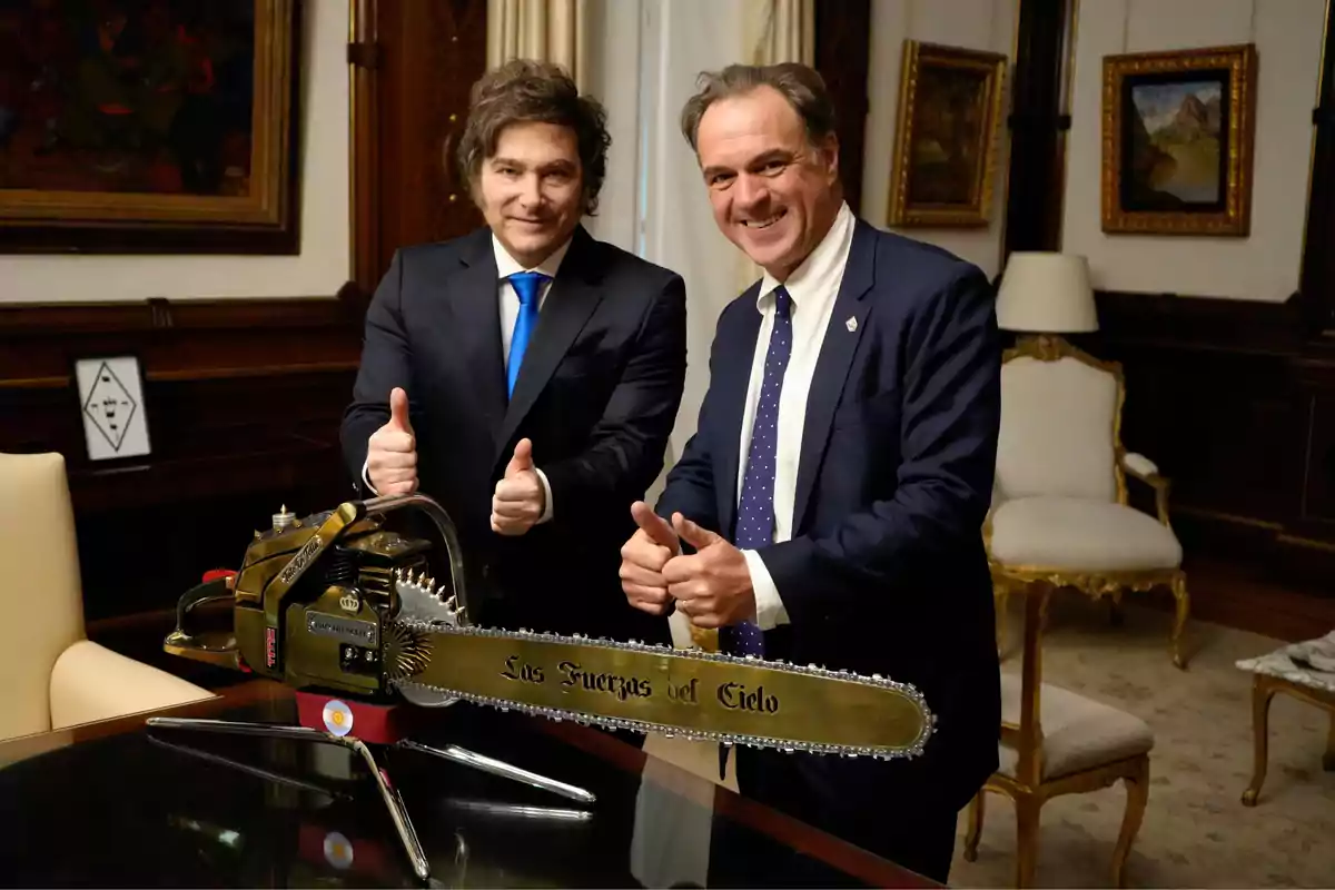 Dos hombres de traje sonríen y levantan los pulgares junto a una motosierra dorada que está apoyada sobre una mesa en una oficina elegante