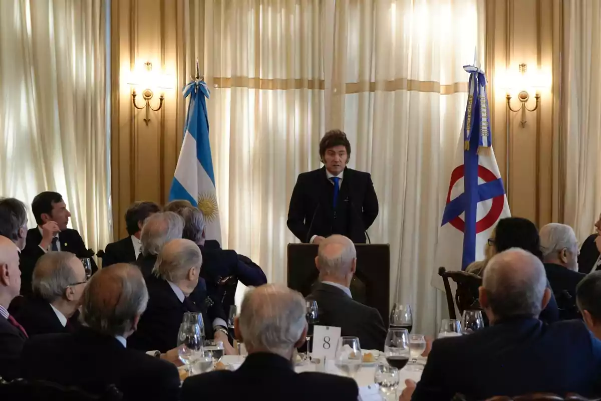 A man in a suit speaks at a podium in front of a group of people seated in an elegant room with flags and light-colored curtains in the background. A man in a suit speaks at a podium in front of a group of people seated in an elegant room with flags and light-colored curtains in the background.