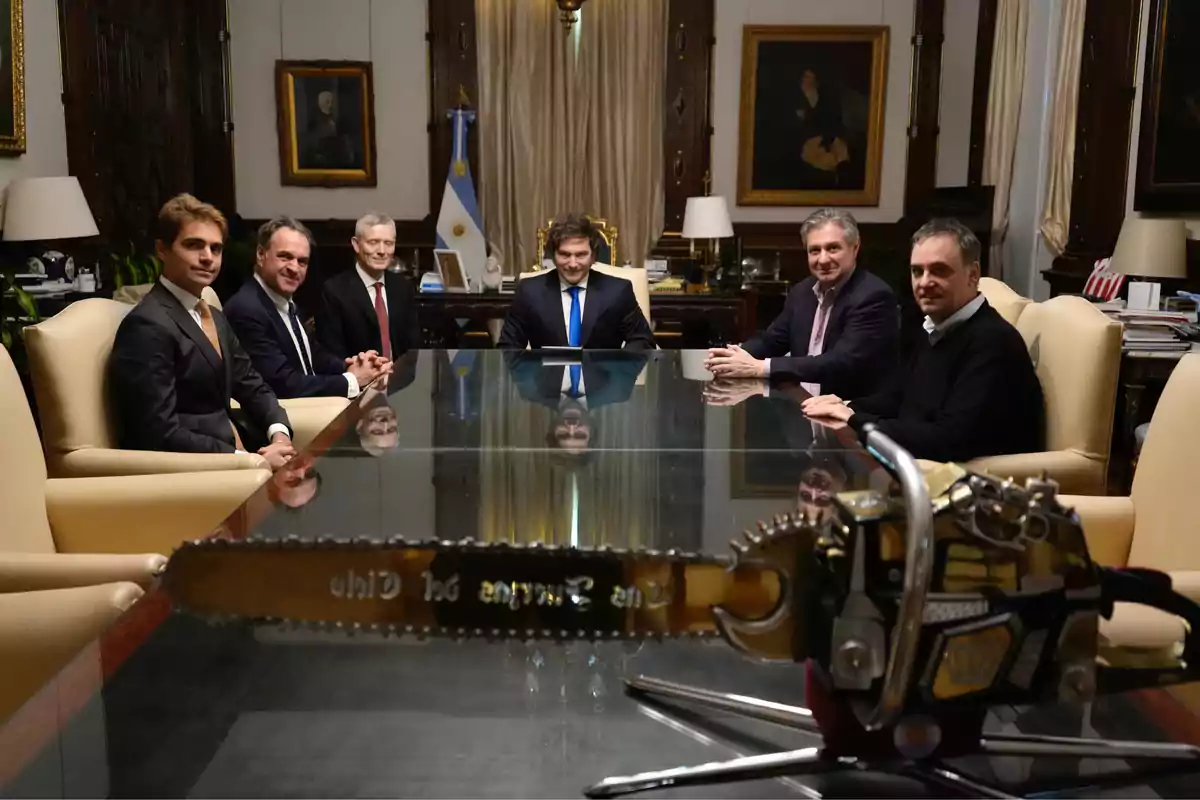 Seis hombres con traje sentados alrededor de una mesa de vidrio en una oficina elegante con una motosierra dorada en primer plano Seis hombres con traje sentados alrededor de una mesa de vidrio en una oficina elegante con una motosierra dorada en primer plano