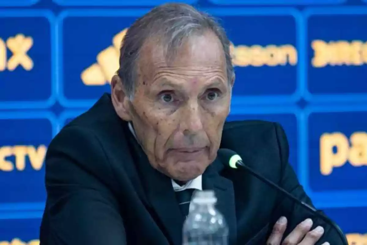 Older man with gray hair and a dark suit speaking at a press conference with a blue background and a microphone in front of him Older man with gray hair and a dark suit speaking at a press conference with a blue background and a microphone in front of him
