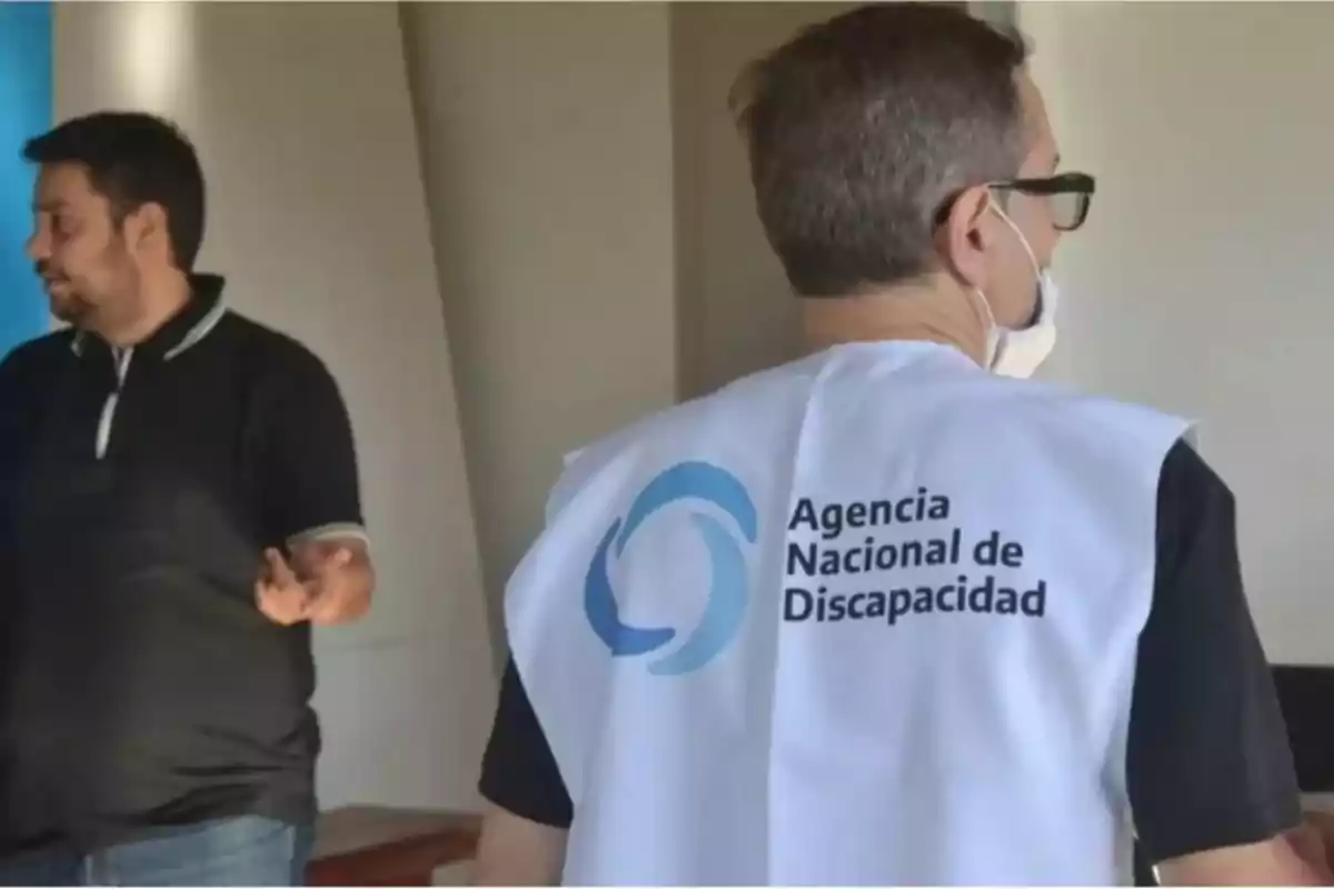 Dos hombres conversan en un interior, uno de ellos lleva un chaleco blanco con el logo y nombre de la Agencia Nacional de Discapacidad.