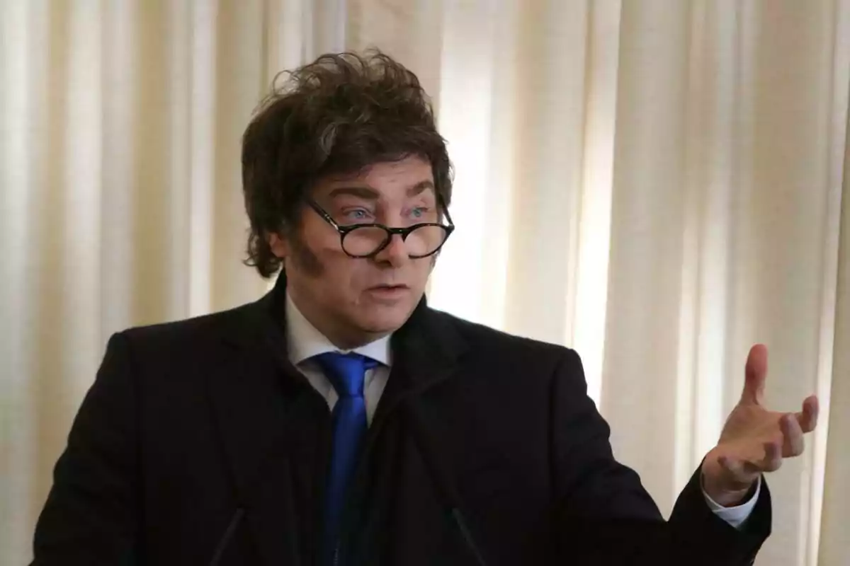 Dark-haired, curly-haired man with glasses and a dark suit speaking and gesturing in front of a light-colored curtain Dark-haired, curly-haired man with glasses and a dark suit speaking and gesturing in front of a light-colored curtain