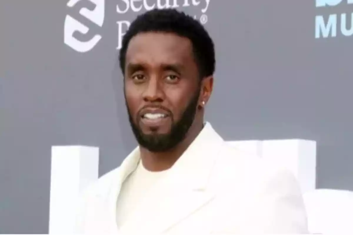 Hombre con barba y cabello corto vestido con traje blanco posando frente a un fondo gris en un evento