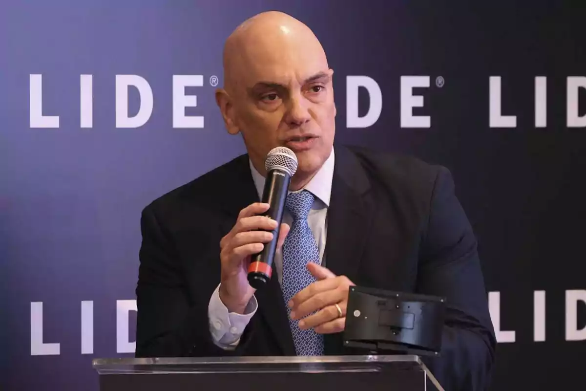 Bald man in a suit and blue tie speaking into a microphone at an event with a background that has the word LIDE repeated. Bald man in a suit and blue tie speaking into a microphone at an event with a background that has the word LIDE repeated.