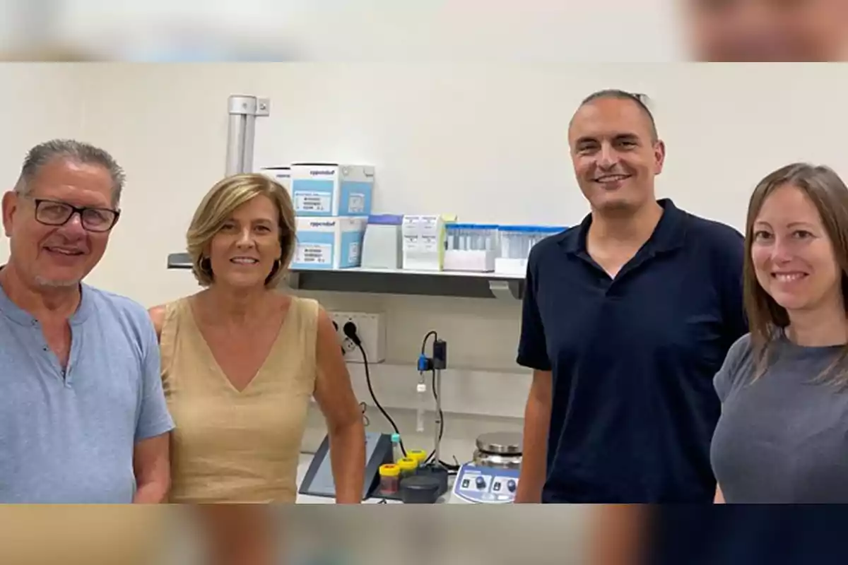 Four people are smiling in a laboratory with scientific equipment and boxes of materials in the background. Four people are smiling in a laboratory with scientific equipment and boxes of materials in the background.