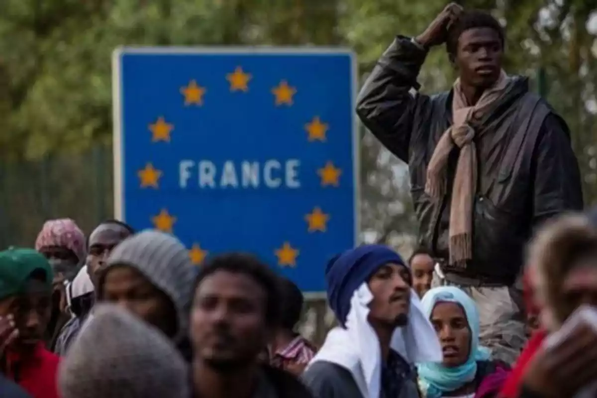Un grupo de personas migrantes se encuentra frente a un cartel azul que indica la entrada a Francia rodeado de estrellas amarillas de la Unión Europea Un grupo de personas migrantes se encuentra frente a un cartel azul que indica la entrada a Francia rodeado de estrellas amarillas de la Unión Europea