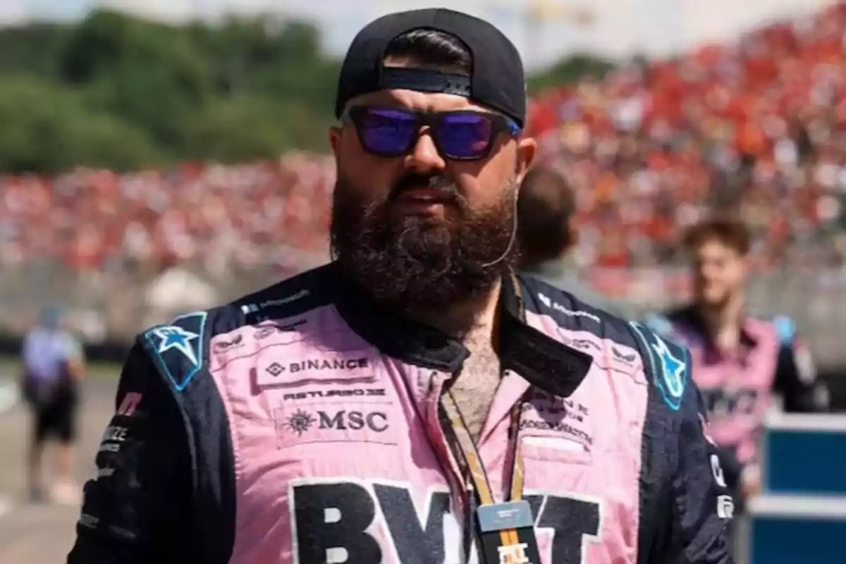 Man with a beard and sunglasses wearing a pink racing uniform and a black backwards cap at an outdoor sporting event