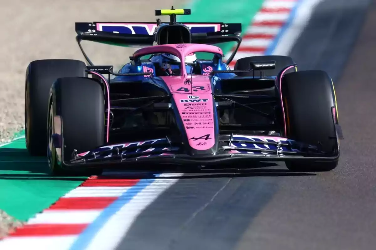 A pink and blue Formula 1 single-seater drives on a track with green, red, and white edges. A pink and blue Formula 1 single-seater drives on a track with green, red, and white edges.