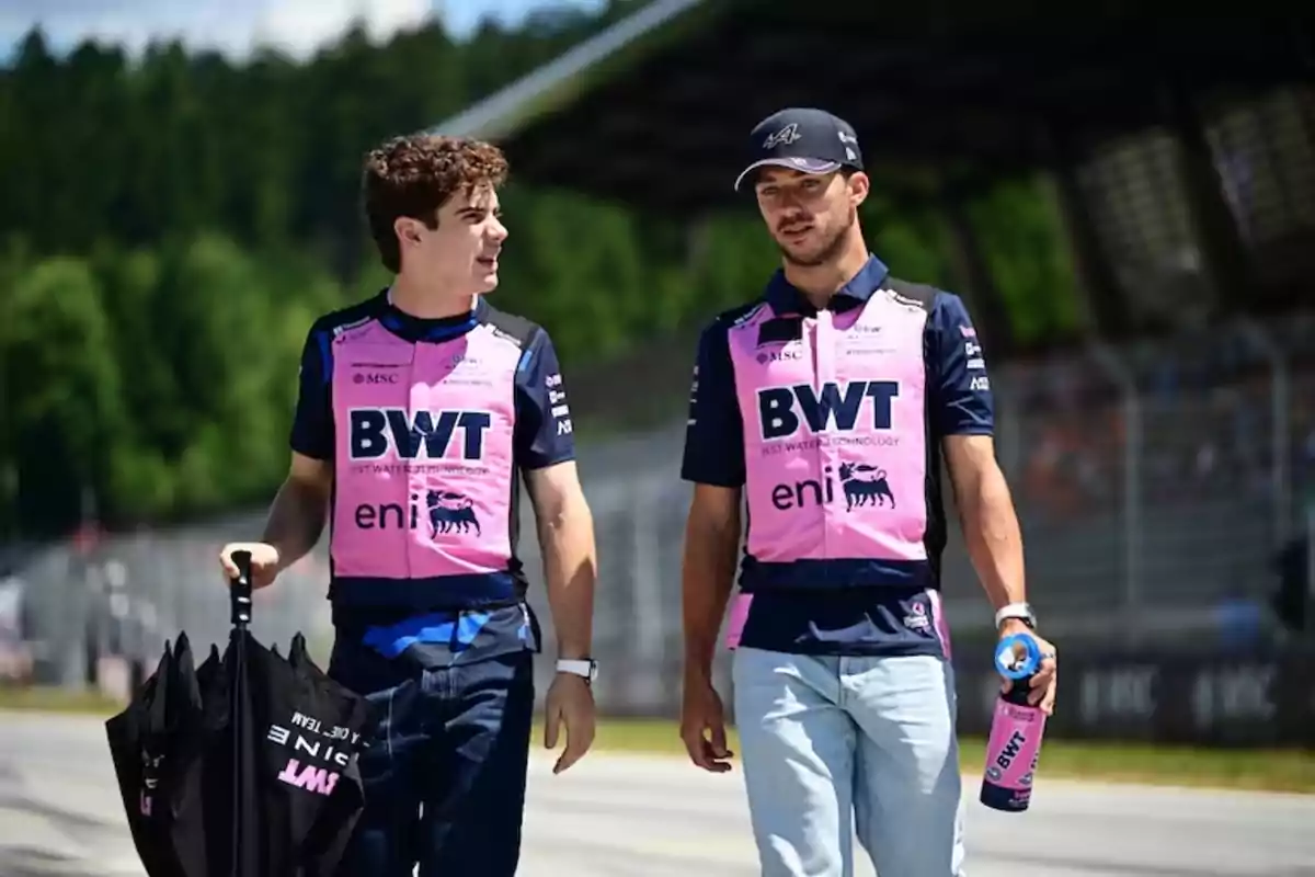 Two men wearing pink vests from a motorsport team walk through the paddock of a racetrack; one is carrying an umbrella and the other a bottle, and they talk as they walk. Two men wearing pink vests from a motorsport team walk through the paddock of a racetrack; one is carrying an umbrella and the other a bottle, and they talk as they walk.