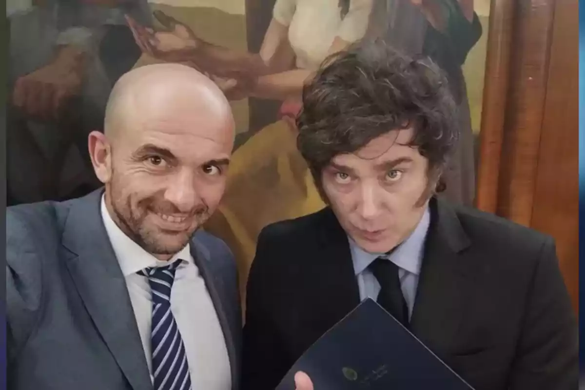 Dos hombres vestidos de traje posan para una selfie en un ambiente formal con una pintura de fondo